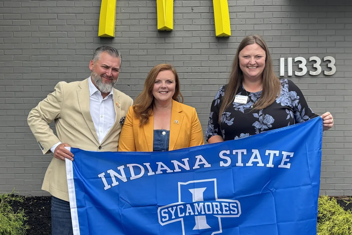An image features three individuals holding a large, blue "Indiana State University Sycamores" flag in front of a grey brick building adorned with a prominent yellow McDonald's logo.