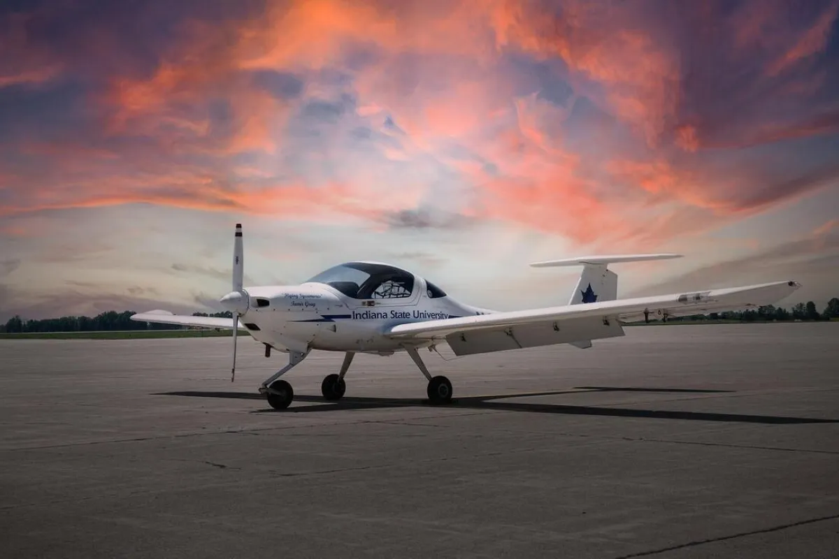 An airplane branded with "Indiana State University" is parked on a runway. The sky in the background features a vibrant sunset with hues of orange, pink, and purple.