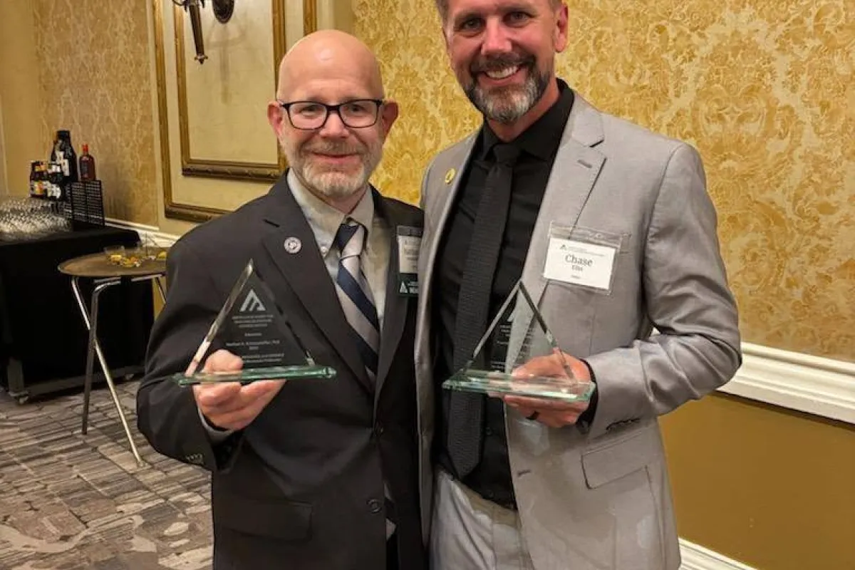Two individuals (Dr. Schaumleffel, left and Chase Ellis, right) are standing in a room with ornate wallpaper, holding clear triangular awards. They are both dressed in formal suits with name tags. 