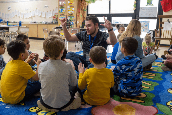 A classroom with 13 young children of mixed races, wearing a variety of yellow, blue, and grey clothing. The children are divided into two groups, sitting in circles on a blue, green, and yellow-colored floor rug. In the left group, a white male sits with the students. He has short brown hair with a matching beard. He wears a black T-shirt and blue jeans. He is holding up both hands as he looks at the students. In the right group, a white female sits with the students.