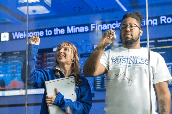 Two students write on a clear dry erase board with news ticker in background