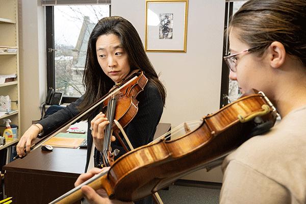 An Asian female professor with black shirt plays a violin while looking at sheet music as a White female student follows along.