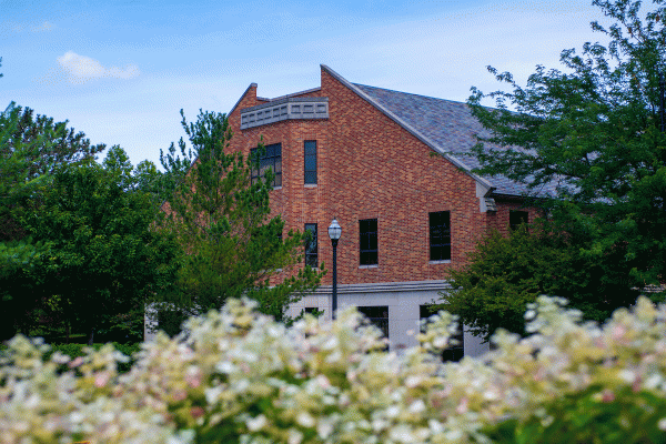 Brick building with a peaked roof partially obscured by trees and flowering shrubs in the foreground. The building’s architecture features tall windows and a decorative roofline, set against a clear, blue sky, creating a peaceful and picturesque campus scene
