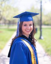 An Honors College student wearing a blue graduation cap and gown, a yellow stole, and a black and blue Honors College hood. The student is standing outside on campus.