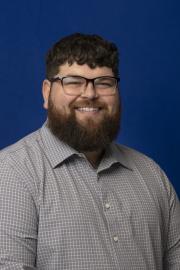A smiling man with short curly hair, glasses, and a full beard, wearing a gray checkered button-down shirt, posed against a solid blue background.