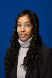 A studio portrait of a woman with long, wavy dark hair wearing a light gray turtleneck and a dark blazer, looking at the camera with a slight smile against a solid blue background.