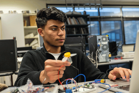 A male student with a dak olive complexion and short black hair sits at a table with a laptop in front of him. He wears a black zipped-up jacket with a white undershirt. Blue and red wiring is visible on the table. He holds a small grey electrical device in his right hand, with a yellow top and grey propellers. Other electrical devices are visible on the table. Computer monitors, a bookcase with stacks of books, and a row of windows are visible in the background. 