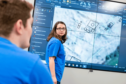Female students with a blue shirt and black framed glasses stands in front of a smart board depicting a satellite map with various areas highlighted.