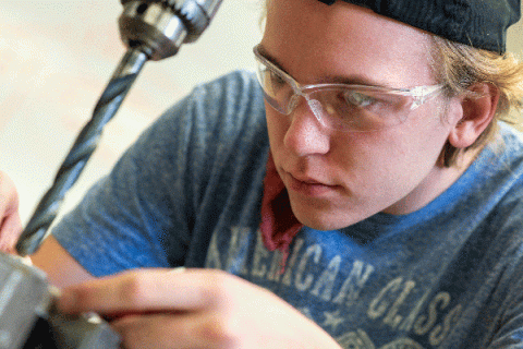 A white male student with blonde hair and wearing a black cap, glasses, and a powder-blue American Classic t-shirt, works on something that is out of focus at bottom left of frame. A drill is visible with a lengthy drill bit that extends down toward whatever he is holding in his fingers. The student’s bright blue eyes are visible as he focuses on his work.  