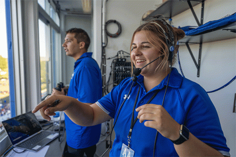 A female student wearing a blue shirt pointing out of the window of a press box with a male student in blue shirt in the background.