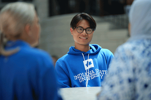 A male student of Asian descent with short dark hair, glasses, and a blue hoodie with Indiana State University in white lettering smiles while sitting at a table. He talks to two female students with their backs to the camera.  