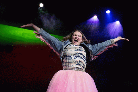 A young white woman with shoulder-length dark brown hair stands on a stage with outstretched arms, wearing a blue jean jacket, a colorful patterned shirt, and a fluffy pink skirt. She smiles excitedly under colorful stage lights.