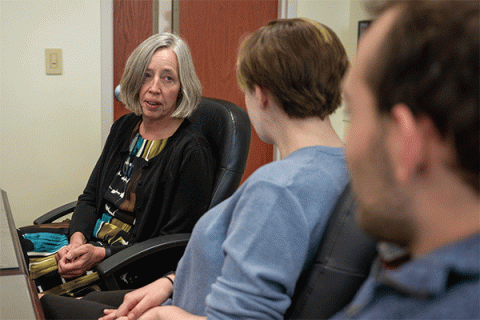 Three individuals sit in black office chairs, engaged in conversation in a room with a wooden door and light-colored walls. An older woman with shoulder-length gray hair, wearing a patterned dress and black sweater, is talking to two younger people facing her. The one nearer her wears a blue top and has short hair, while the other is partly visible, with short, dark brown hair and a beard. 
