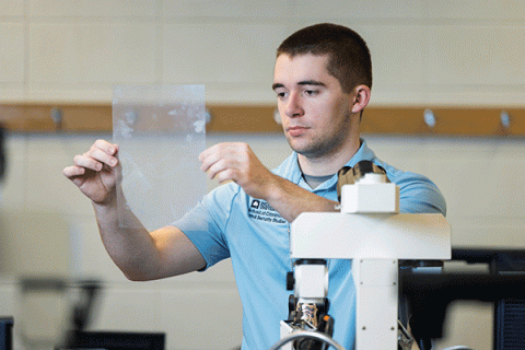 A male student with short, dark hair, and wearing a light blue Indiana State University shirt, examines a transparent sheet in a forensic science lab, with specialized lab equipment visible in the foreground.