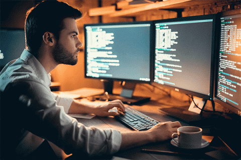 A young man with dark hair and a short beard, wearing a white shirt with folded sleeves, sits in front of a computer with three monitors on it. He is typing. The room is dimly lit, and a cup of coffee sits on the desk beside him.