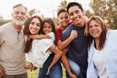 Six people of different generational age groups stand together outdoors. They are smiling. The group includes one older man and woman, one younger man and woman, and one boy and girl. The children are carried on the backs of the two young adults. Trees and greenery are visible in the background.