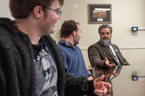 Three male individuals are seated around a table in a meeting room. The person on the left is wearing glasses, a black jacket, and a T-shirt with an image of a historical figure. The person in the middle is wearing a blue sweater. The person on the right has gray hair and a beard, and is dressed in a brown suit with a striped tie. A framed picture is on the white wall behind them. 