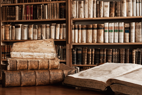 A library scene with shelves filled with old, leather-bound books in the background. In the foreground, there is a wooden table with several ancient books stacked on top of each other and one large book open, displaying its aged pages. The overall atmosphere suggests a historical or scholarly setting.