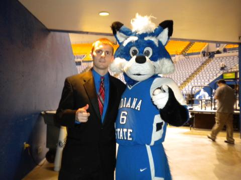 A White man in a black suit and red tie poses beside a blue-and-white Indiana State University mascot wearing a basketball uniform. They stand in an indoor arena with yellow and blue seats visible in the background.