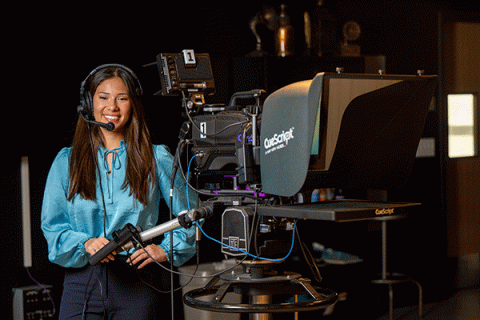 A smiling young woman with long, dark brown hair and wearing a headset with a microphone stands beside a professional video camera setup in a studio. The camera includes a teleprompter labeled "CueScript." The woman is smiling, and the background features shelves and studio equipment, indicating a broadcast or recording environment.