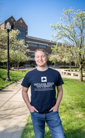 A while male wearing a navy blue Indiana State University T-shirt stands on a sunny campus walkway, with green lawns, blooming trees, and a brick academic building in the background.