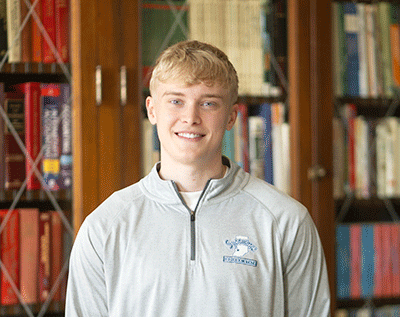 “A young man smiling while standing indoors in front of bookshelves, wearing a light gray quarter-zip pullover.”