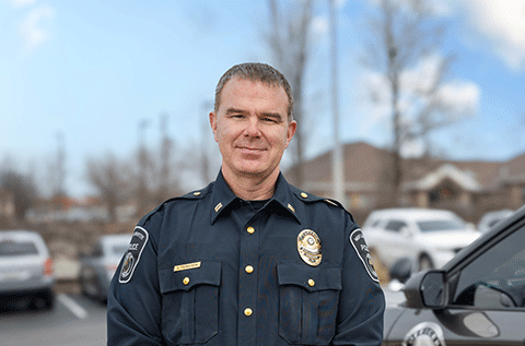 “Portrait of a man wearing a police uniform, standing outdoors with vehicles visible in the background.”