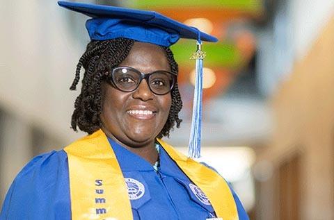 "Person wearing a blue graduation cap and gown with a yellow stole that reads “Summa,” smiling in a brightly lit hallway with colorful ceiling decorations."