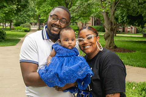 “Two adults holding a baby outdoors, smiling, with trees and a walkway in the background.”