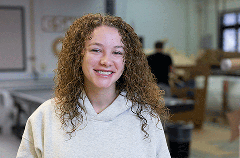 “Portrait of a woman with curly hair wearing a light-colored hoodie, smiling indoors with a workshop setting in the background.”