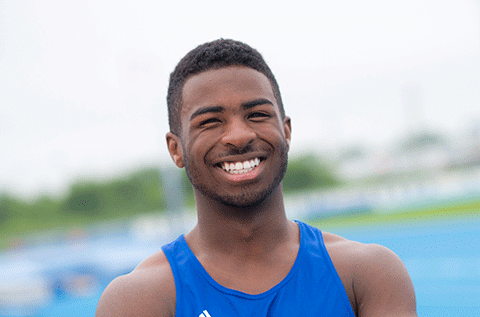 “Portrait of a man wearing a blue athletic top, smiling outdoors with a sports setting in the background.”