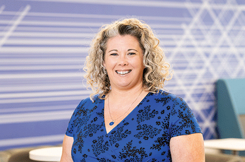 “Portrait of a woman with curly hair wearing a blue patterned top, smiling indoors with a graphic background.”