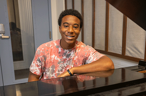 “Portrait of a man wearing a patterned shirt, seated indoors near a piano, smiling toward the camera.”