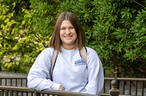 “Portrait of a woman wearing a light-colored sweatshirt and backpack, standing outdoors with greenery in the background.”
