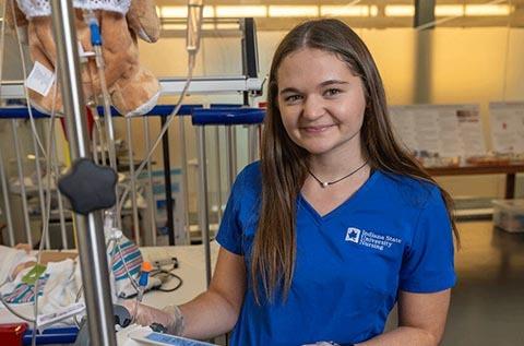 "Person wearing a blue “Indiana State University Nursing” shirt standing in a clinical setting, with medical equipment and a stuffed animal hanging from an IV pole in the background."