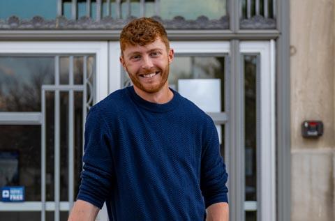 Person standing in front of a building entrance, wearing a dark long‑sleeve shirt, with short curly hair. Glass doors and a keypad entry system are visible in the background.