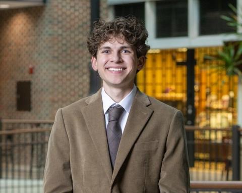 A white male with curly brown hair wearing a brown blazer, white shirt, and tie stands indoors near a railing, with brick walls, large windows, and potted plants in the background, suggesting an atrium.
