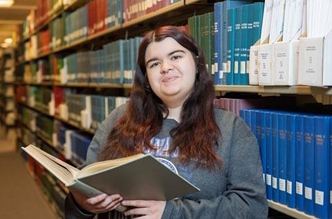 Person standing in a library aisle, holding an open book and looking toward the camera, with shelves of books lining both sides.