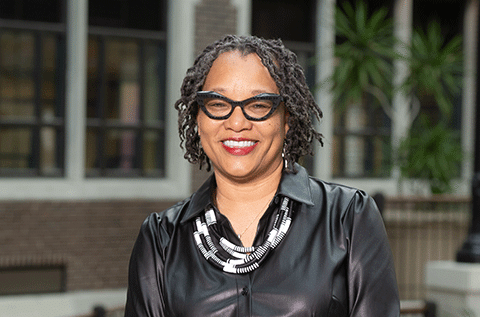 “Portrait of a woman wearing glasses and a black top, smiling indoors, with a building and plants in the background.”