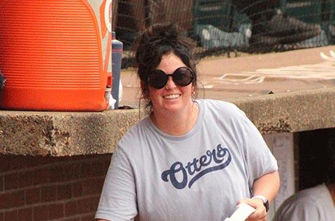 "Person wearing large round sunglasses and a light gray T‑shirt that reads “Otters,” smiling and holding a rolled paper, with an orange water cooler and stadium seating visible in the background."
