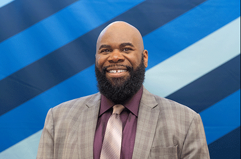 “Portrait of a man wearing a suit and tie, smiling, against a blue patterned background.”