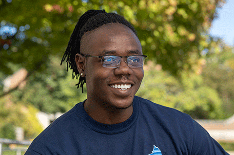 “Portrait of a man wearing glasses and a dark shirt, smiling outdoors with trees in the background.”