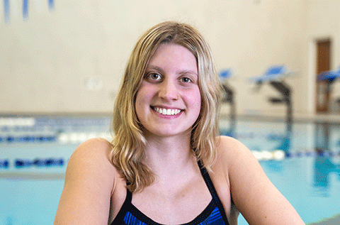 Portrait of a woman wearing a swim top, smiling indoors with a swimming pool in the background.”