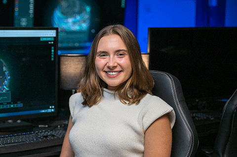 “Portrait of a woman wearing a light-colored top, seated indoors with computer monitors visible in the background.”