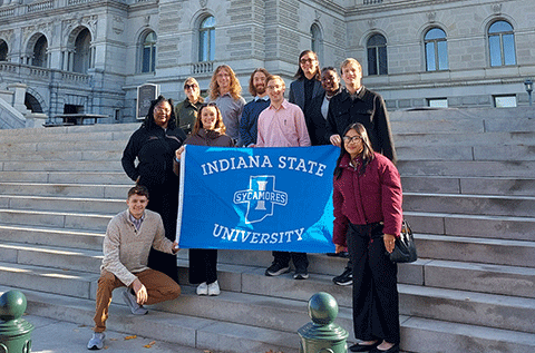 “A group of students and staff standing on outdoor stone steps in front of a large historic building, holding a blue Indiana State University Sycamores banner and smiling for a group photo.”