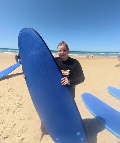A white female student in a wetsuit stands on a sandy beach holding a blue surfboard, with ocean waves, other surfers, and a clear blue sky in the background.
