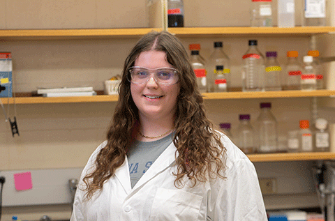 White female student with curly brown hair wearing a white lab coat stands in a laboratory, with shelves of labeled bottles, glassware, and research supplies visible in the background.