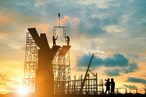 Silhouetted construction workers assemble a large Y shaped structural element at sunset atop scaffolding at sunset, guided by a crane, while two additional workers observe from the ground against a cloudy sky. 