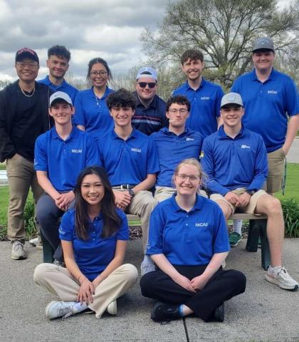 Group of 12 students wearing matching blue polo shirts pose outdoors on and around a bench, with trees and a cloudy sky in the background.