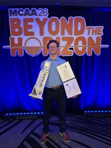 Male standing in front of a large “MCAA 26 Beyond the Horizon” event sign, holding a first-place student chapter competition trophy and certificates, with blue stage lighting in the background.